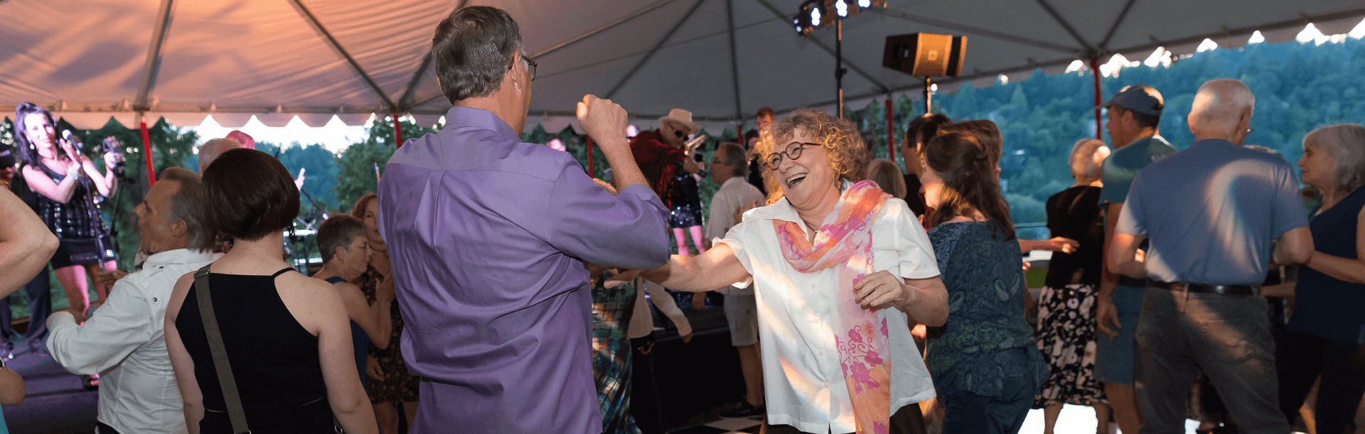 Craig and Rikki are dancing under an outdoor event tent with live music in the background and a crowd of people surrounding them