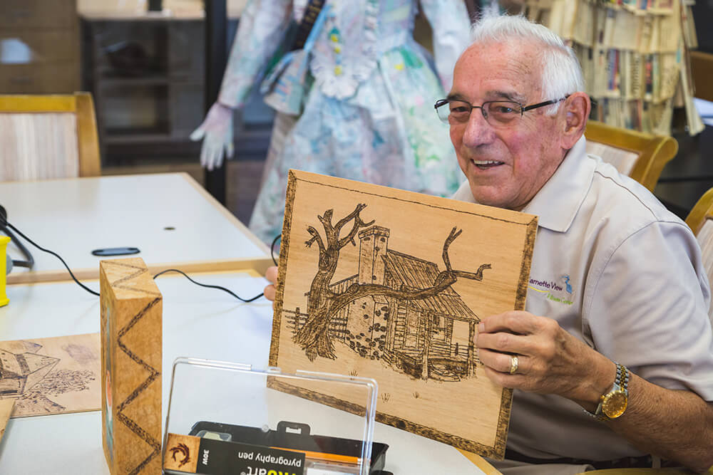 A man holding a piece of art he made by burning a log cabin and a tree into a wood panel