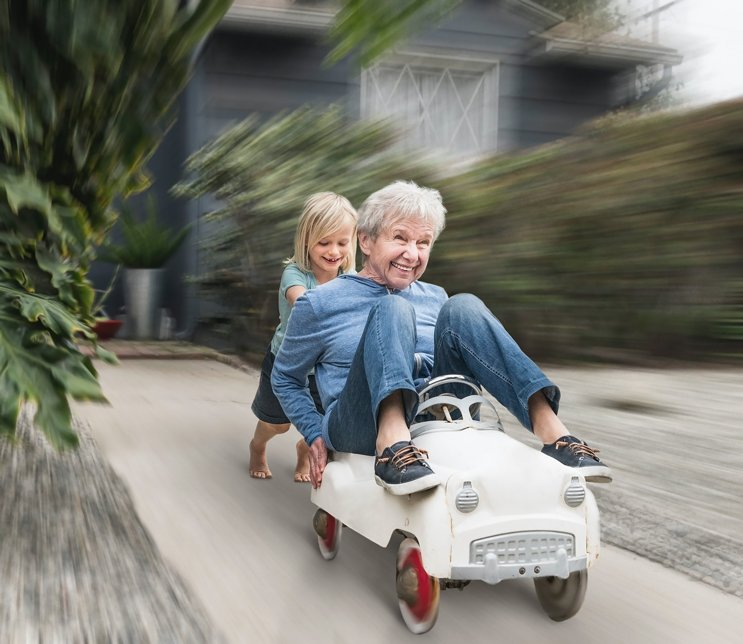 A grandchild pushing her grandmother down the driveway in a toy car
