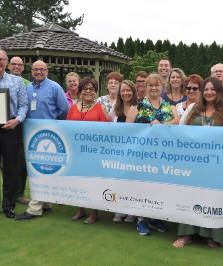 A group of Willamette View staff with a banner in front of them and a plaque signifying becoming a Blue Zone project